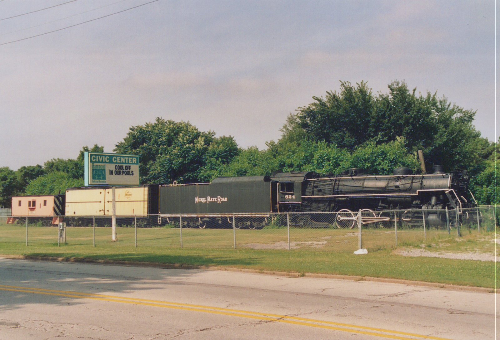 A black steam locomotive numbered 624, labeled Nickel Plate Road, sits on display behind a chain-link fence next to a road, with green trees and a sign for Hammond Civic Center in the background.