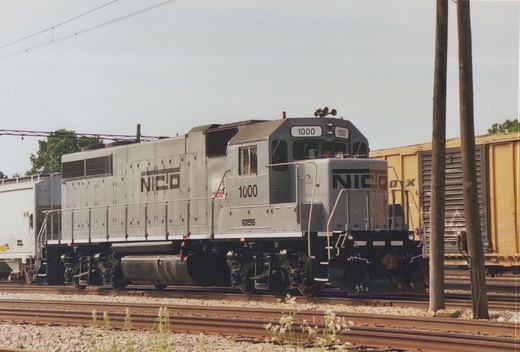 A gray NICTD locomotive numbered 1000 is seen on a railroad track, with freight cars behind it and overhead wires above; one crew member is visible in the cab.