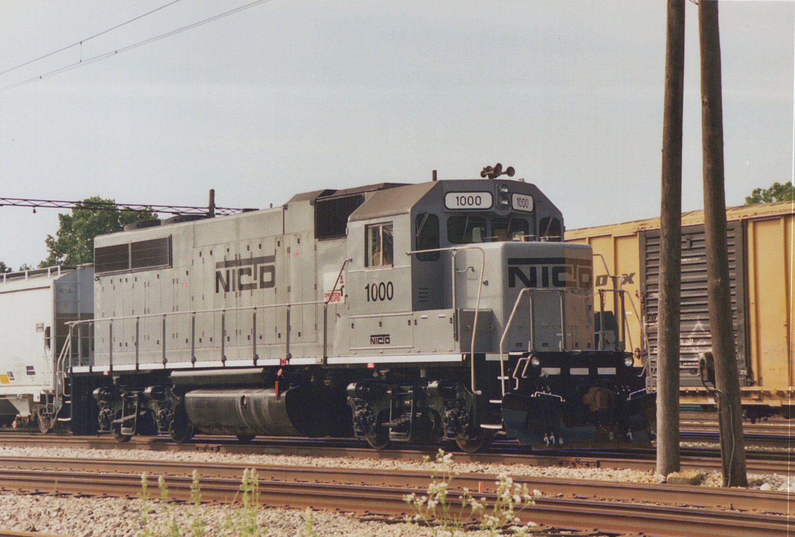 A gray NICTD locomotive numbered 1000 is seen on a railroad track, with freight cars behind it and overhead wires above; one crew member is visible in the cab.