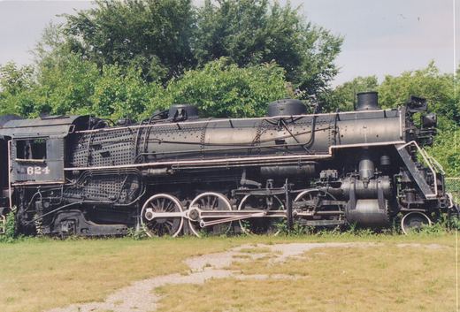 Side view of a black steam locomotive numbered 624, on display in a grassy, tree-lined area under a clear sky.