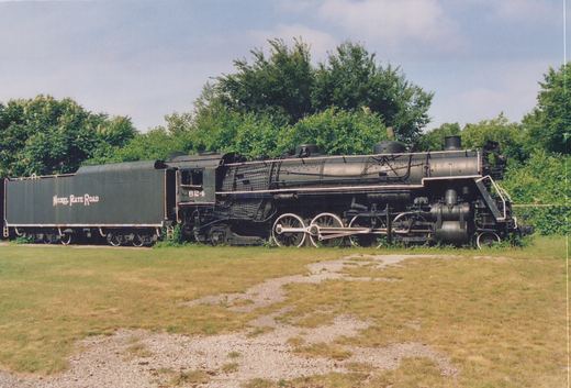 A side view of a black Nickel Plate Road steam locomotive numbered 624, coupled to its tender, sitting on a grassy area with trees behind it.