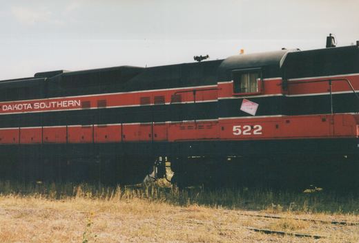 A diesel locomotive with 'Dakota Southern' lettering and the number 522 on its side, standing on a railway track beside dry grass under a clear sky.