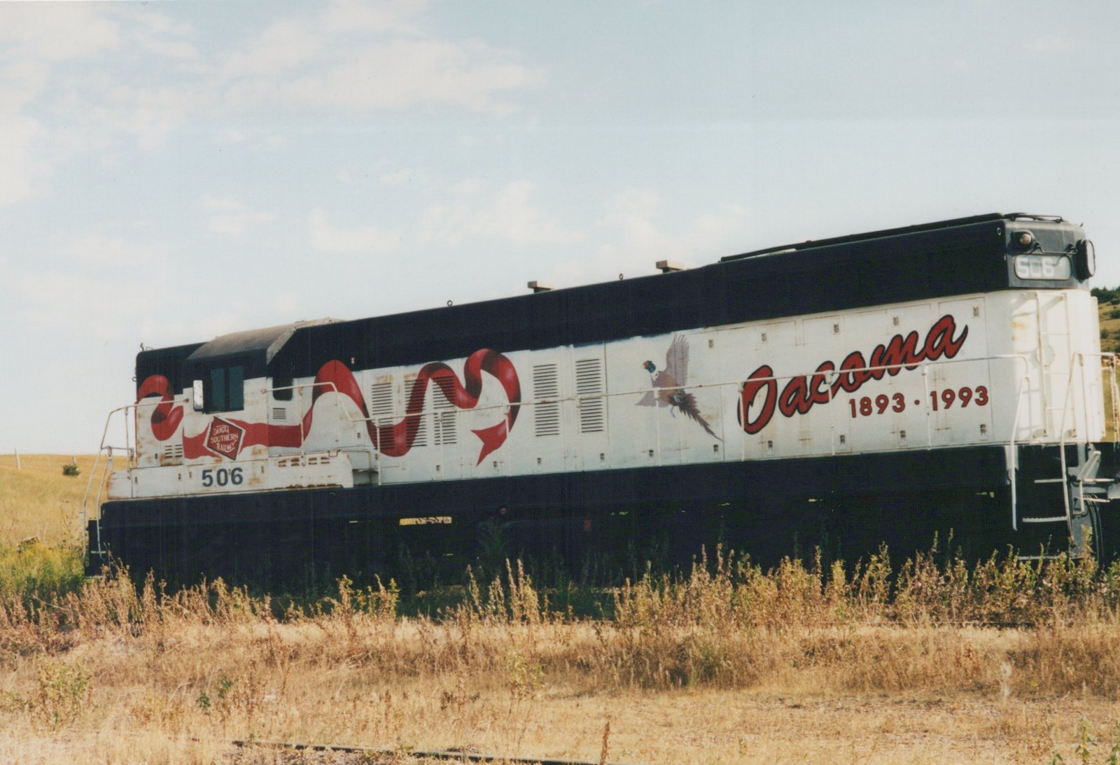 A white and black diesel locomotive with number 506, decorated with red ribbon graphics and a pheasant, and lettered 'Oacoma 1893-1993', stands in a grassy field.