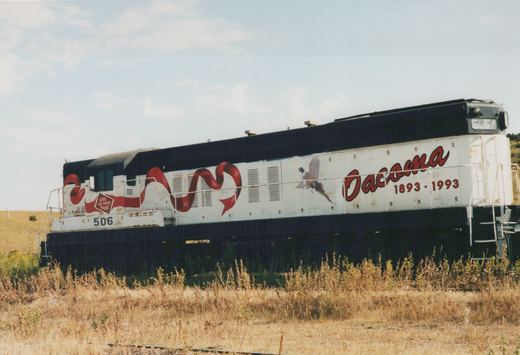 A white and black diesel locomotive with number 506, decorated with red ribbon graphics and a pheasant, and lettered 'Oacoma 1893-1993', stands in a grassy field.