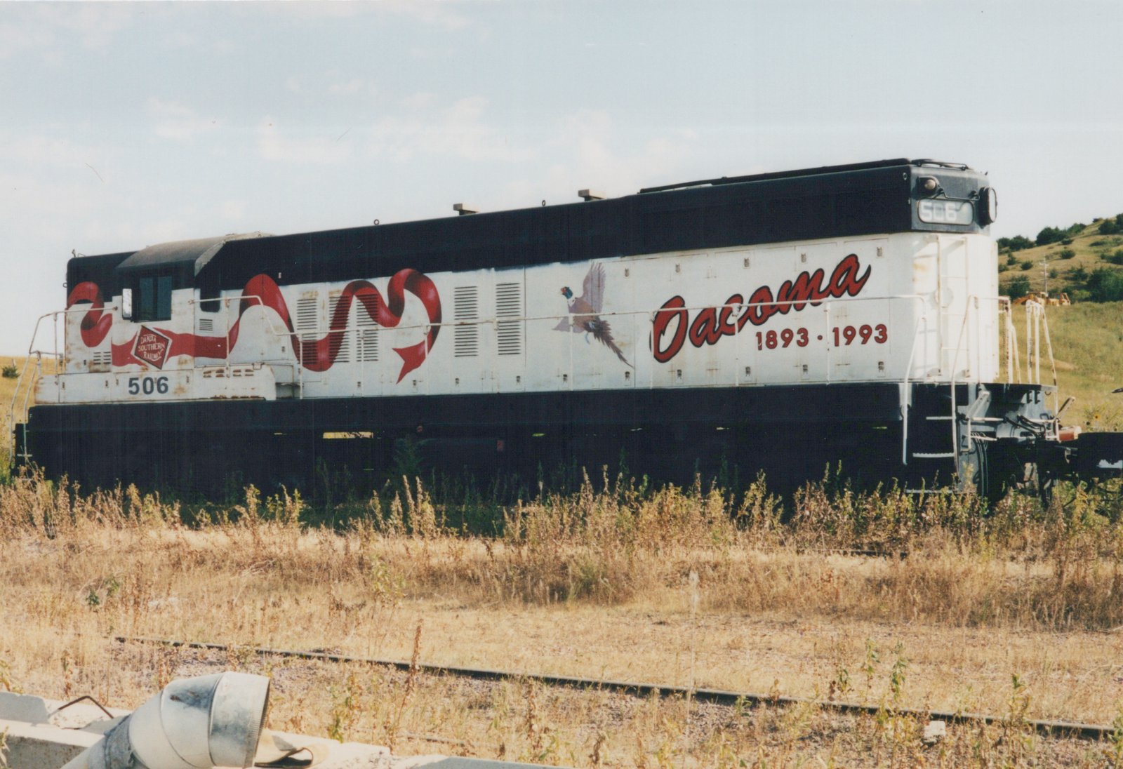 A side view of a white and black diesel locomotive with the number 506 and 'Oacoma 1893-1993' painted on its side, standing on overgrown tracks in a sunny, rural setting.