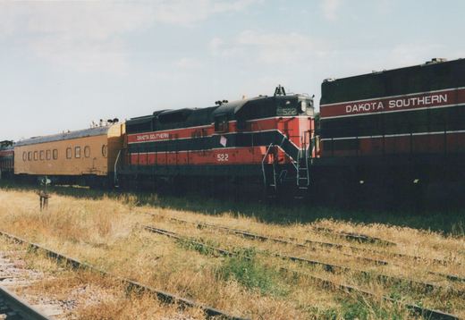A red and black diesel locomotive numbered 522 pulls a yellow passenger car and another locomotive on a set of tracks in a grassy yard under a clear sky.