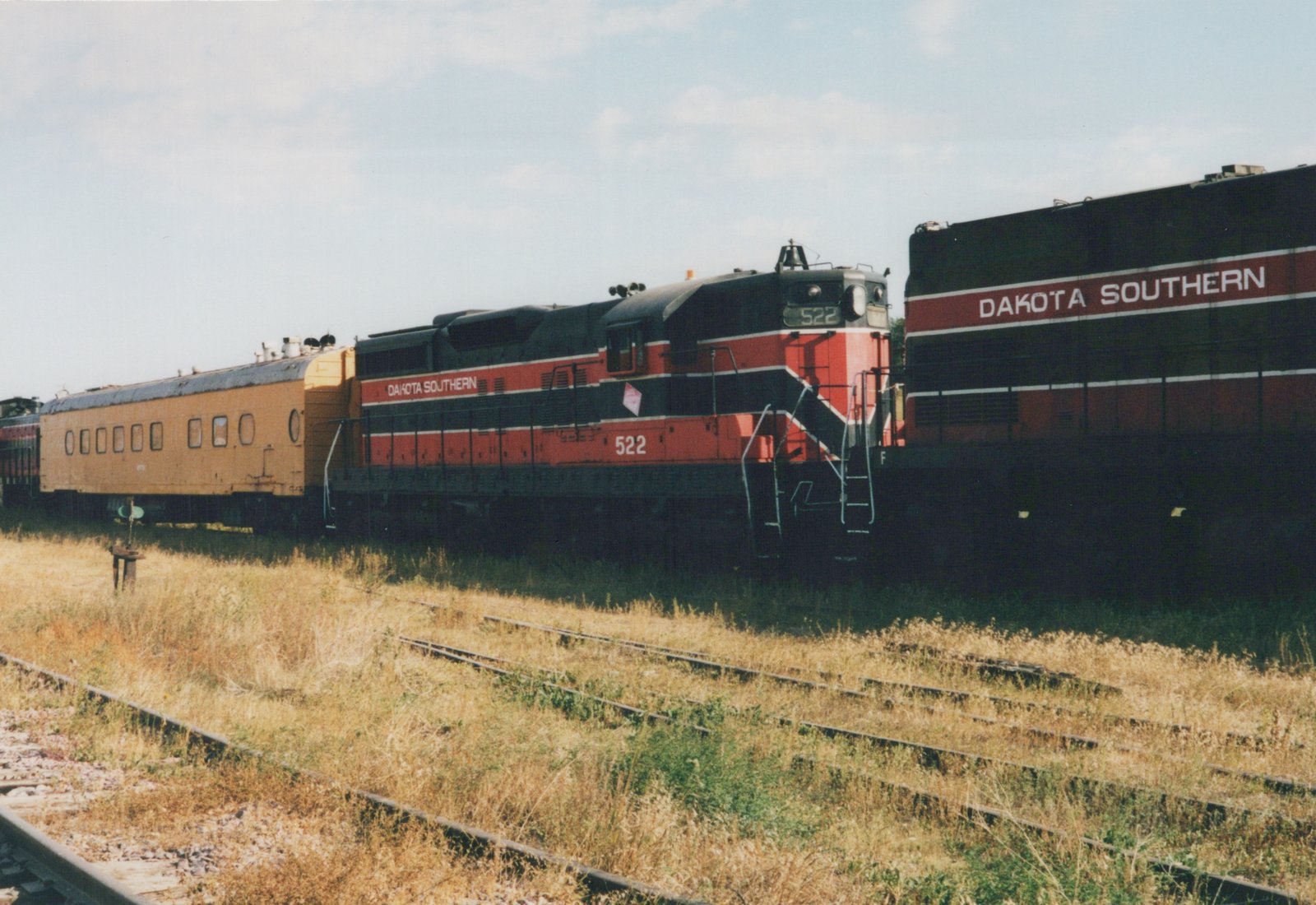 A red and black diesel locomotive numbered 522 pulls a yellow passenger car and another locomotive on a set of tracks in a grassy yard under a clear sky.
