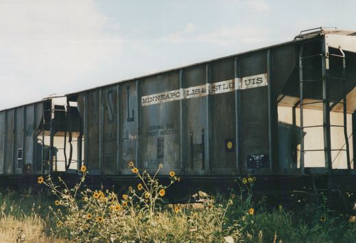 A side view of a dark gray Minneapolis and St. Louis Railway freight car, number 1049, parked on a track next to a field of yellow sunflowers under a clear sky.