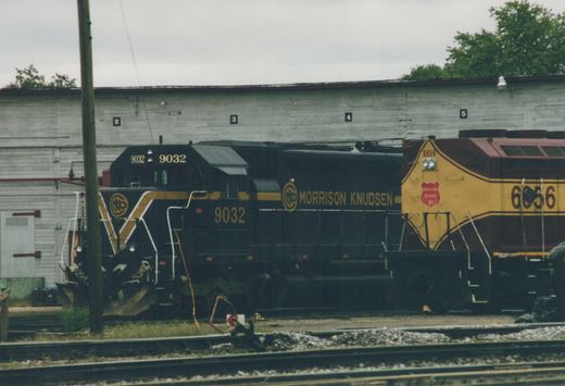 Two diesel locomotives, one black and yellow labeled Morrison Knudsen 9032, and one yellow and maroon labeled Wisconsin Central Ltd 6056, are parked on adjacent tracks in front of a white warehouse.