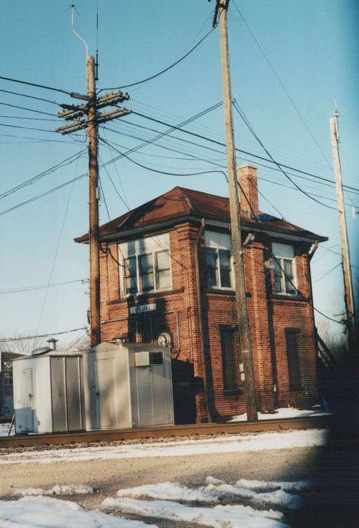 A two-story brick building with a 'DEVAL' sign stands next to a railroad track with snow on the ground and utility poles nearby.