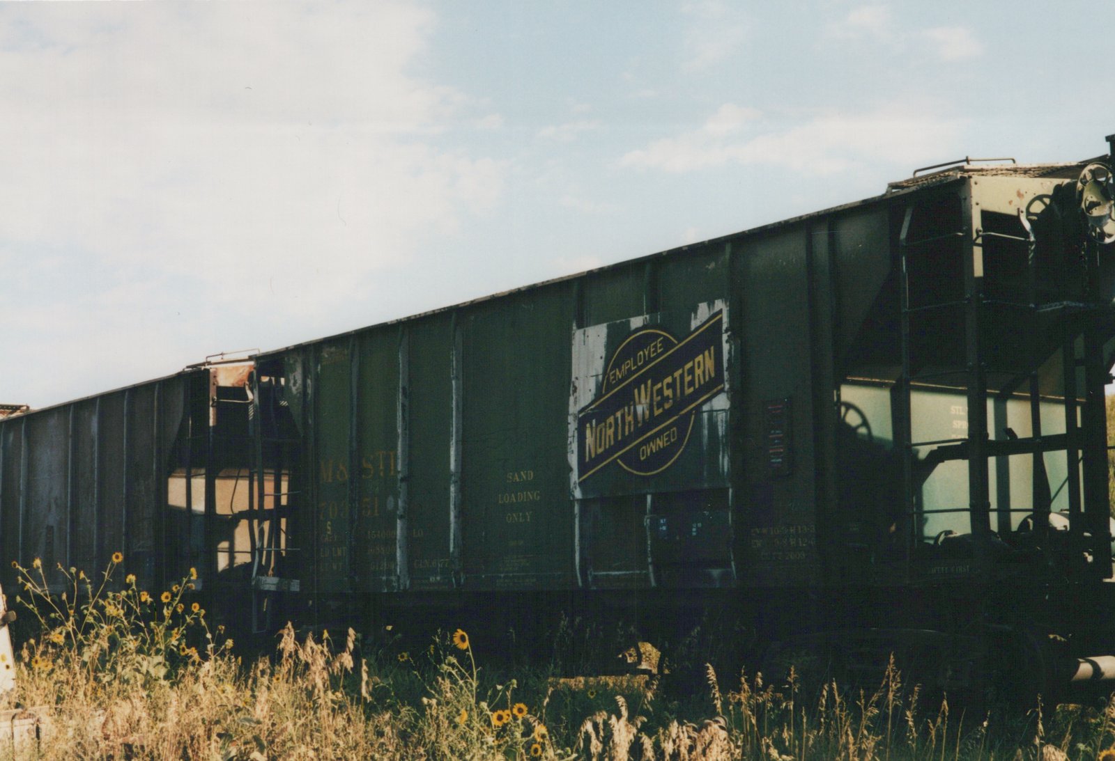 Side view of a dark green M&S STI freight car, number 70351, featuring an 'EMPLOYEE OWNED NORTHWESTERN' herald, parked in a weedy yard.