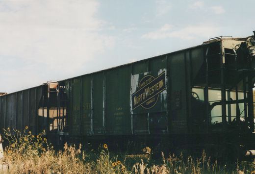 Side view of a dark green M&S STI freight car, number 70351, featuring an 'EMPLOYEE OWNED NORTHWESTERN' herald, parked in a weedy yard.