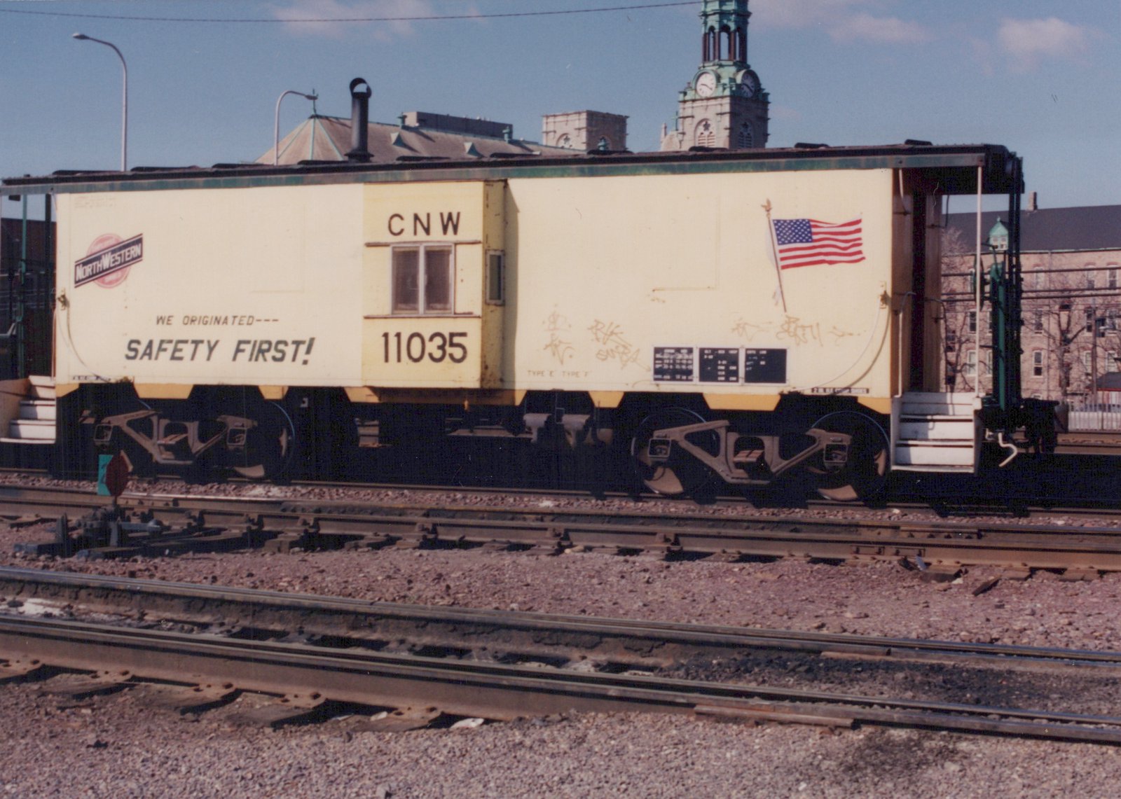 A yellow Chicago and North Western caboose numbered 11035 sits on tracks in a rail yard, with a clock tower and buildings in the background under a clear sky.