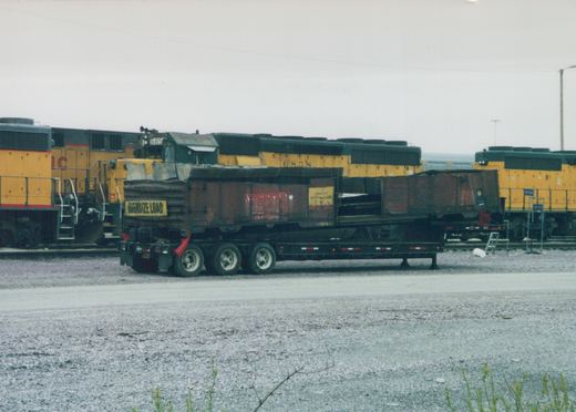 A yellow and green Illinois Central locomotive marked 685 is parked in a gravel yard next to another locomotive, with a black truck trailer carrying an oversize load in front of them under an overcast sky.