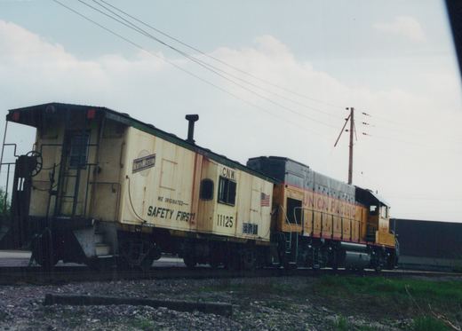 A yellow and green Chicago and North Western caboose marked 11125 is coupled to a yellow Union Pacific locomotive marked 733, both on a gravel track under a cloudy sky.