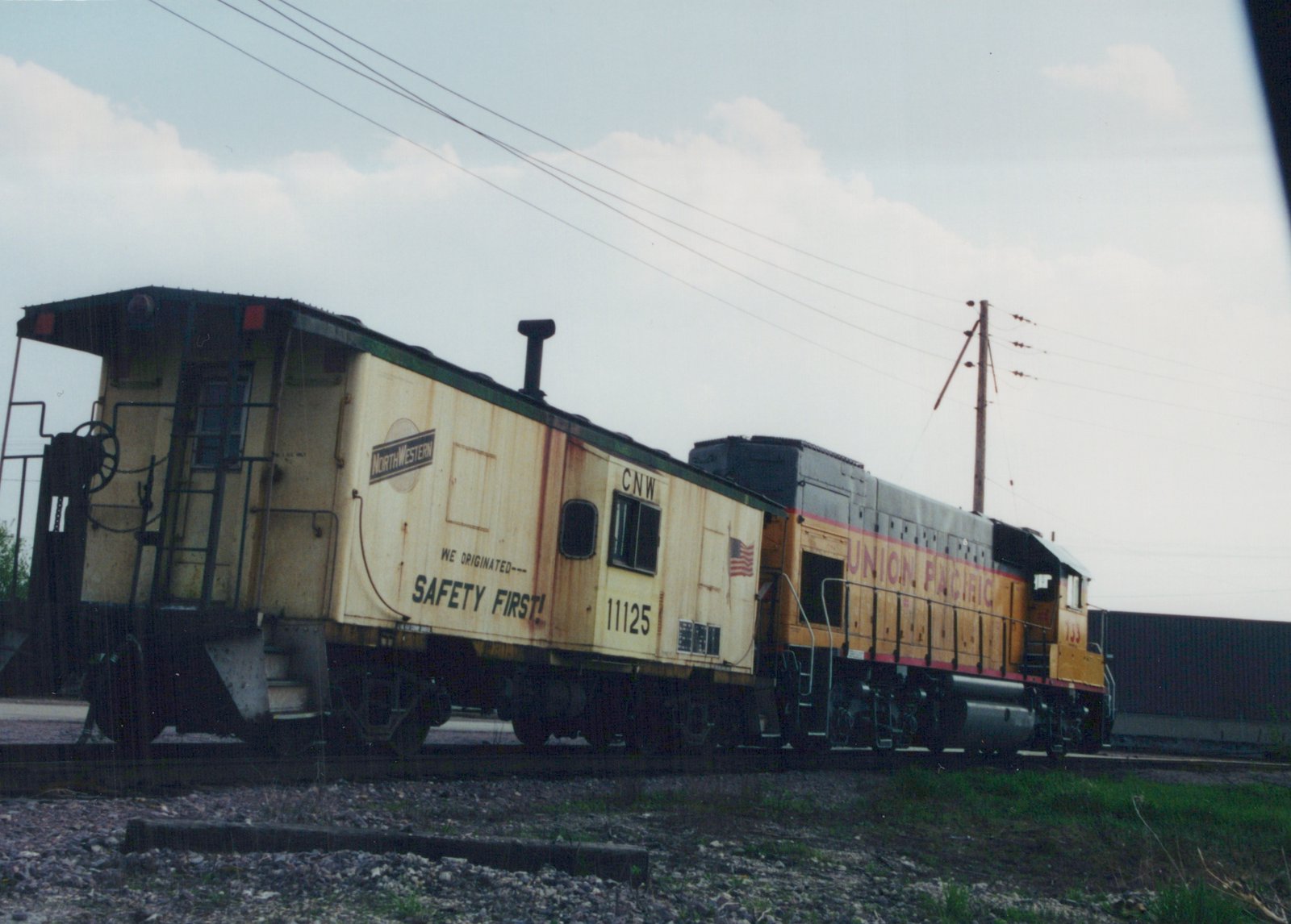 A yellow and green Chicago and North Western caboose marked 11125 is coupled to a yellow Union Pacific locomotive marked 733, both on a gravel track under a cloudy sky.