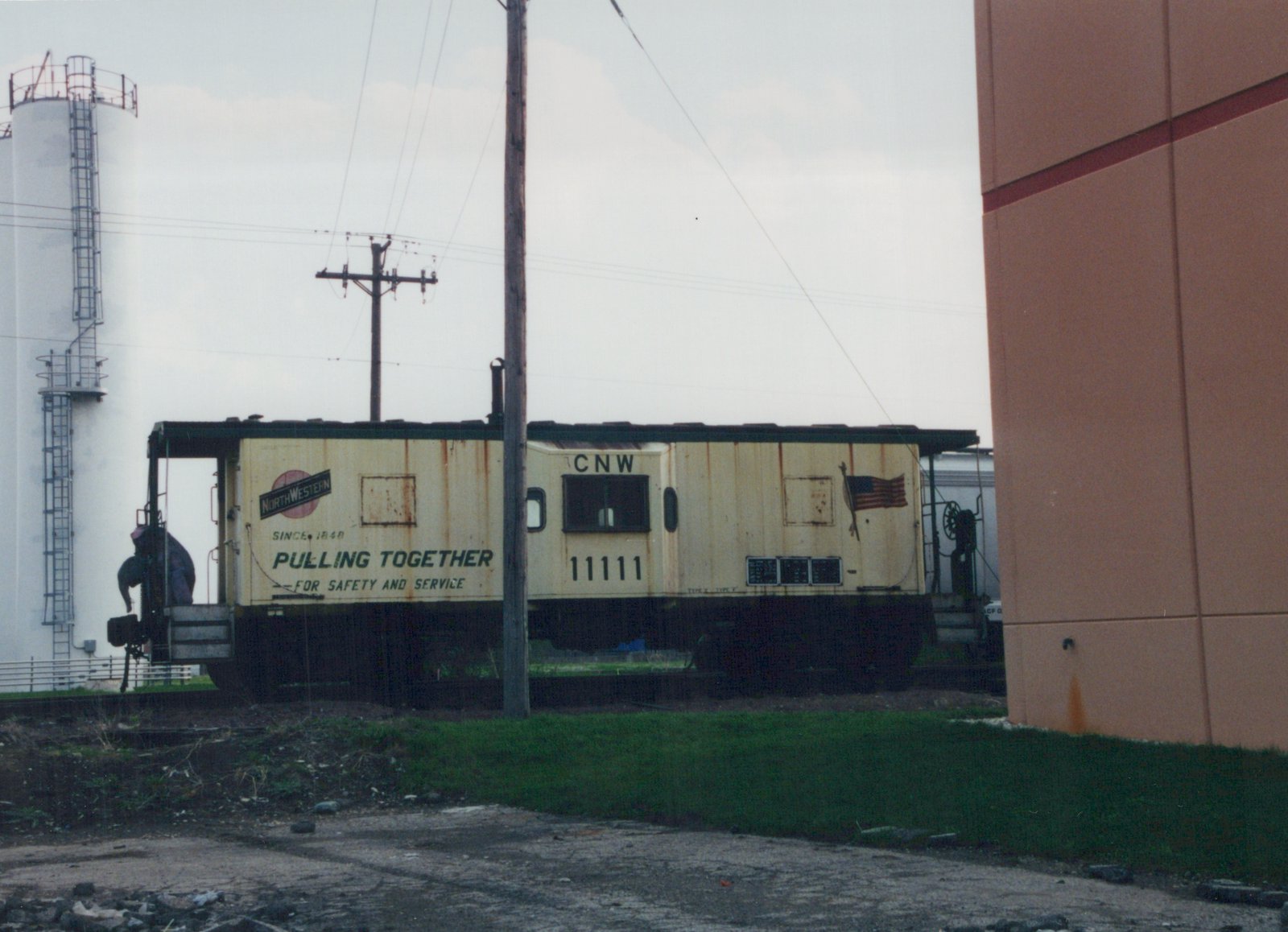 A cream and green Chicago and North Western caboose numbered 11111 is parked on a track in a rail yard, with a person standing on its rear platform.