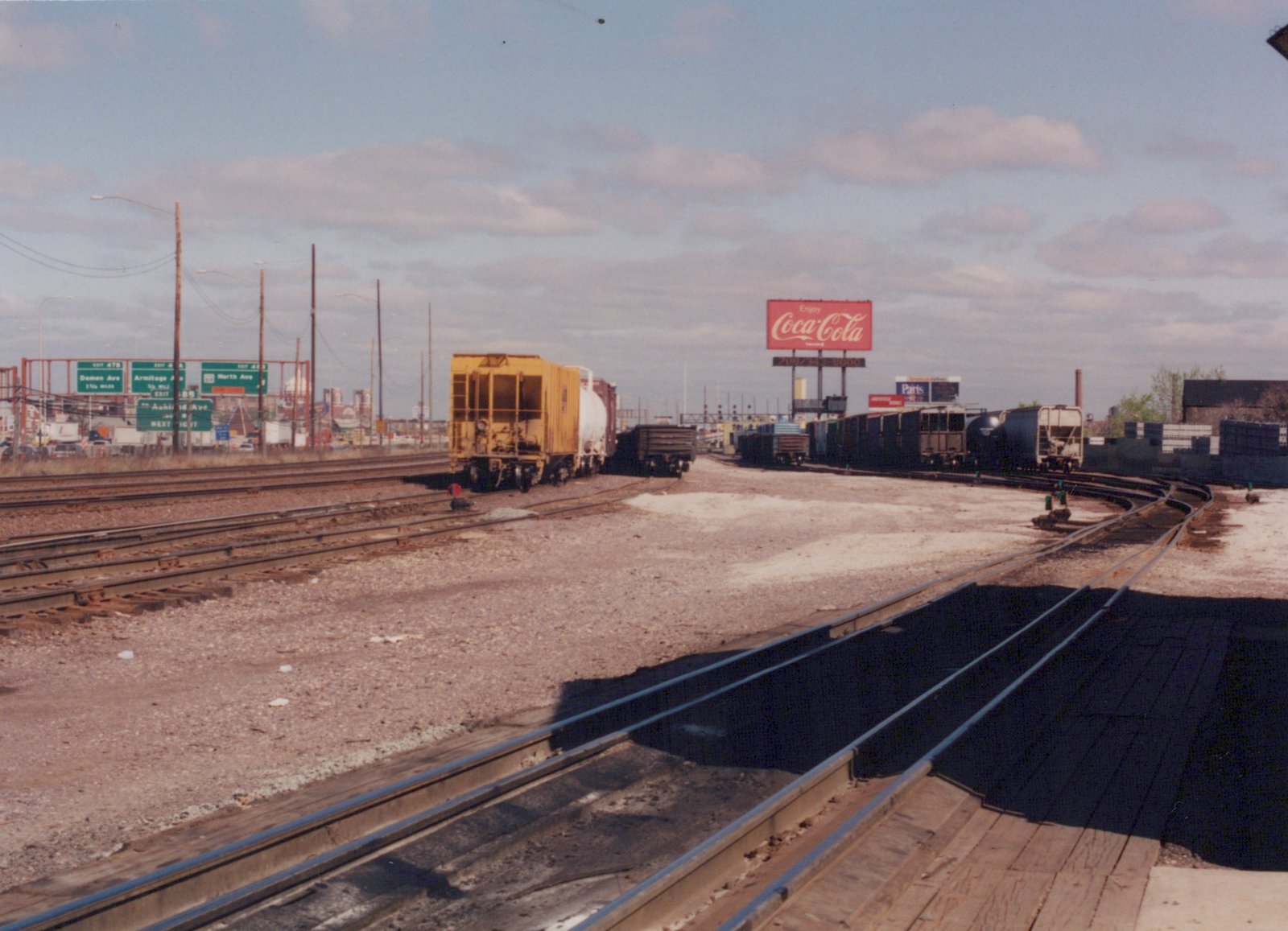 A view of a freight rail yard with several tracks, a variety of freight cars including a yellow caboose and a white tank car, a large red Coca-Cola billboard, and green highway signs in the distance.