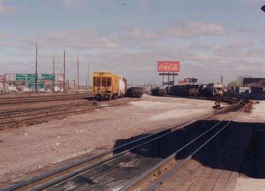 A view of a freight rail yard with several tracks, a variety of freight cars including a yellow caboose and a white tank car, a large red Coca-Cola billboard, and green highway signs in the distance.