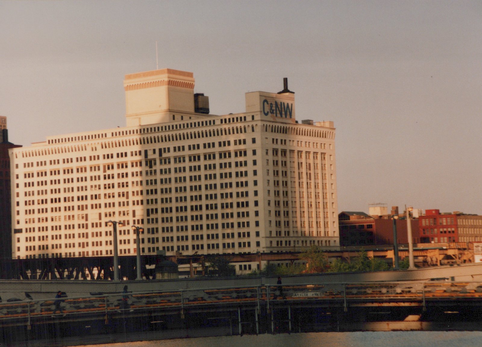 A large building with 'C&NW' lettering on its side stands behind an elevated railway, with people walking on a bridge below.