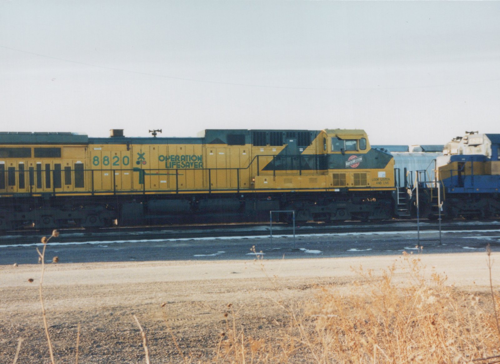 A yellow and black diesel locomotive with the number 8820 and 'Operation Lifesaver' lettering on its side, belonging to the Chicago and North Western Railway, is parked in a yard with patches of snow on the ground.