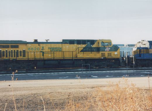 A yellow and black diesel locomotive with the number 8820 and 'Operation Lifesaver' lettering on its side, belonging to the Chicago and North Western Railway, is parked in a yard with patches of snow on the ground.