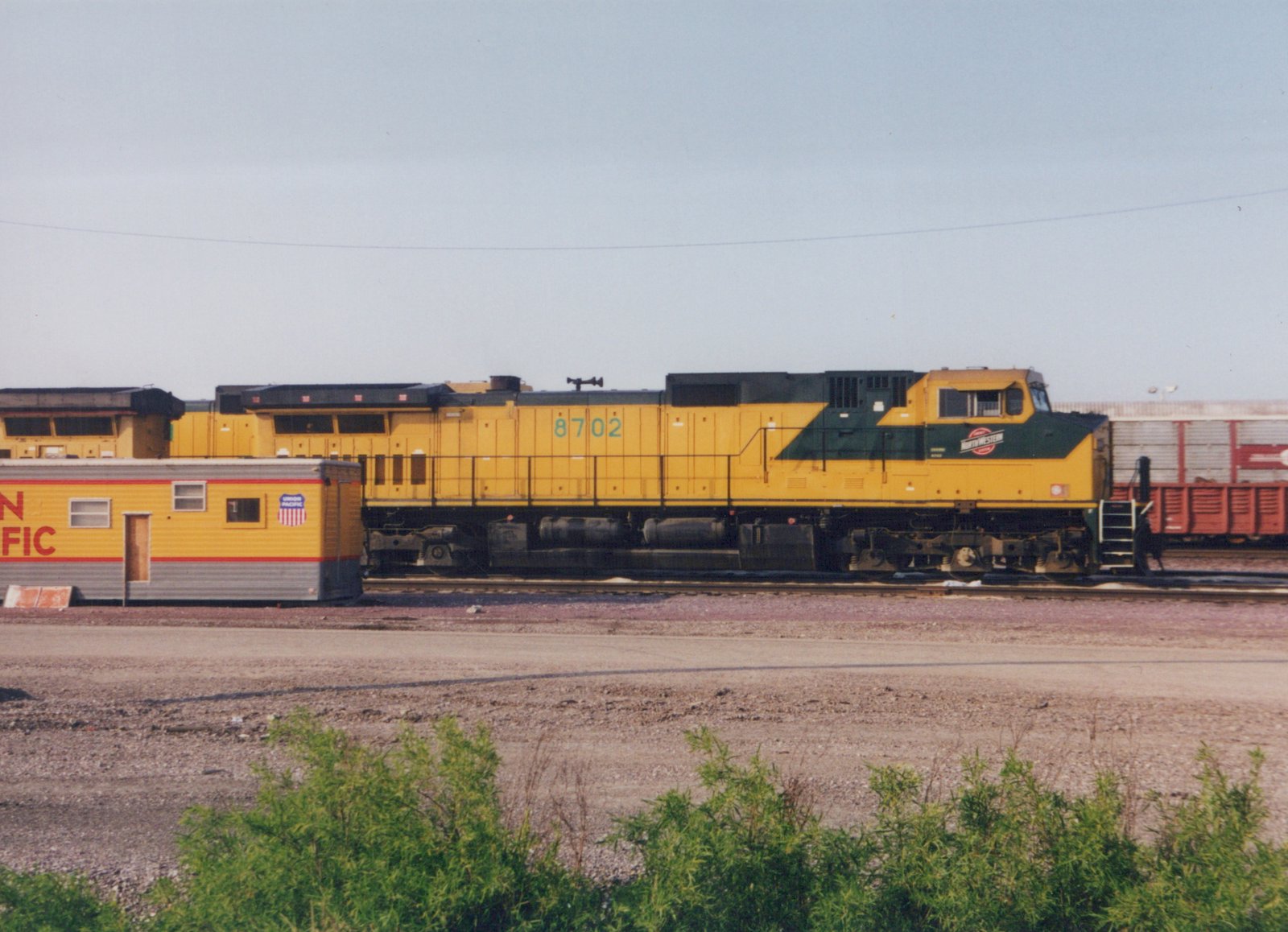 A yellow and green diesel locomotive with the number 8702 is parked in a rail yard next to a yellow office car with Union Pacific markings.