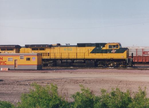 A yellow and green diesel locomotive with the number 8702 is parked in a rail yard next to a yellow office car with Union Pacific markings.