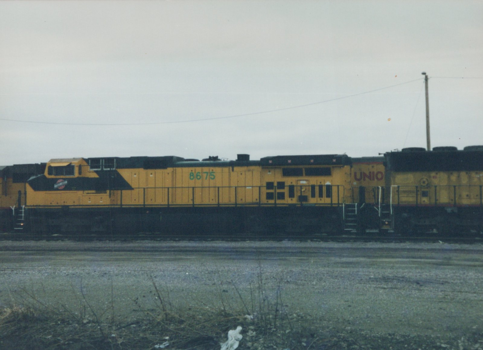 A yellow and black diesel locomotive with the number 8675 and Chicago and North Western lettering sits on a track in a gravel yard under an overcast sky.