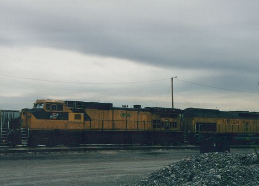 A side-view photograph of a Chicago and North Western locomotive numbered 8688 in orange and black, paired with a Union Pacific locomotive, in a gravel yard under a cloudy sky.