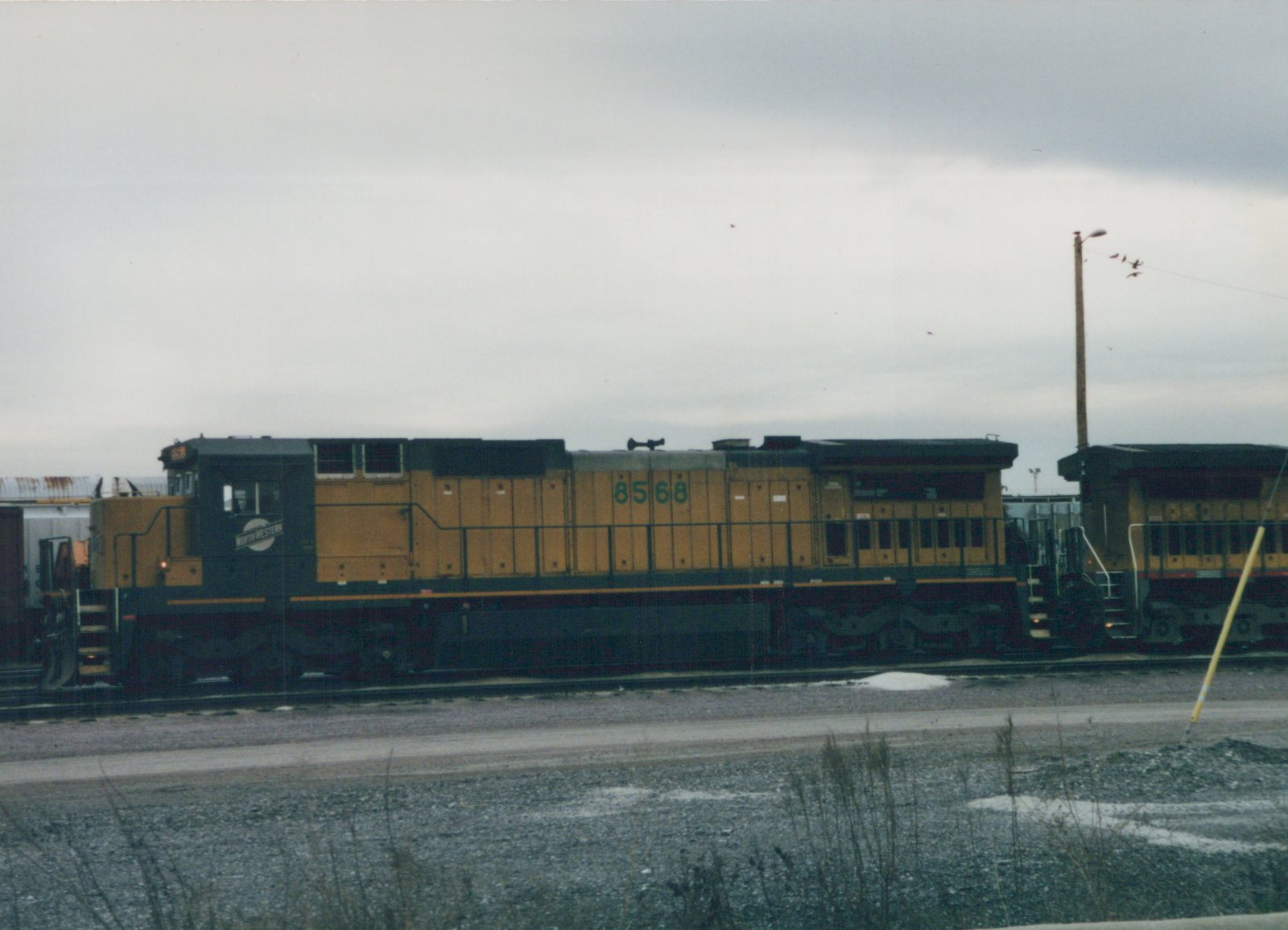 A yellow and dark green diesel locomotive with the number 8568 and 'NORTHWESTERN' on its side sits on railroad tracks in a gravel yard with snow patches under a gray sky.