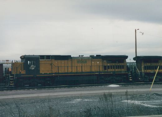 A yellow and dark green diesel locomotive with the number 8568 and 'NORTHWESTERN' on its side sits on railroad tracks in a gravel yard with snow patches under a gray sky.