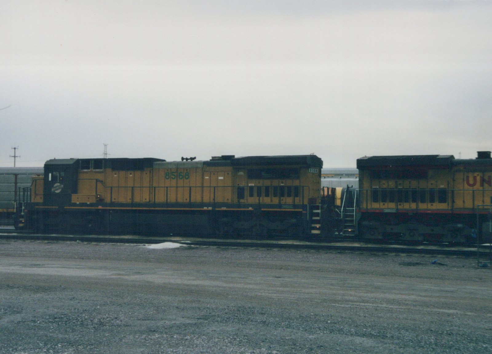 Side view of two diesel locomotives on parallel tracks in a gravel yard, one Chicago and North Western number 8568, the other Union Pacific, under a gray sky with snow patches on the ground.