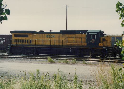 A side view of a yellow and black Chicago and North Western diesel locomotive, number 8524, sitting on tracks in a rail yard with weeds and a utility pole in the background.