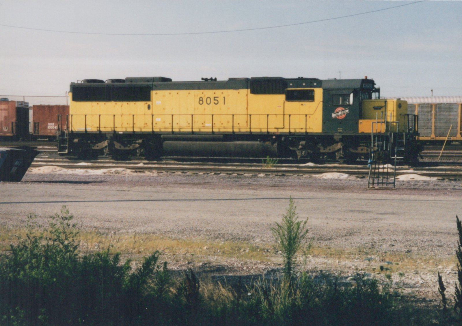 A side view of a yellow and green Chicago and North Western locomotive numbered 8051, parked on tracks in a railyard with freight cars in the background.