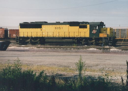 A side view of a yellow and green Chicago and North Western locomotive numbered 8051, parked on tracks in a railyard with freight cars in the background.