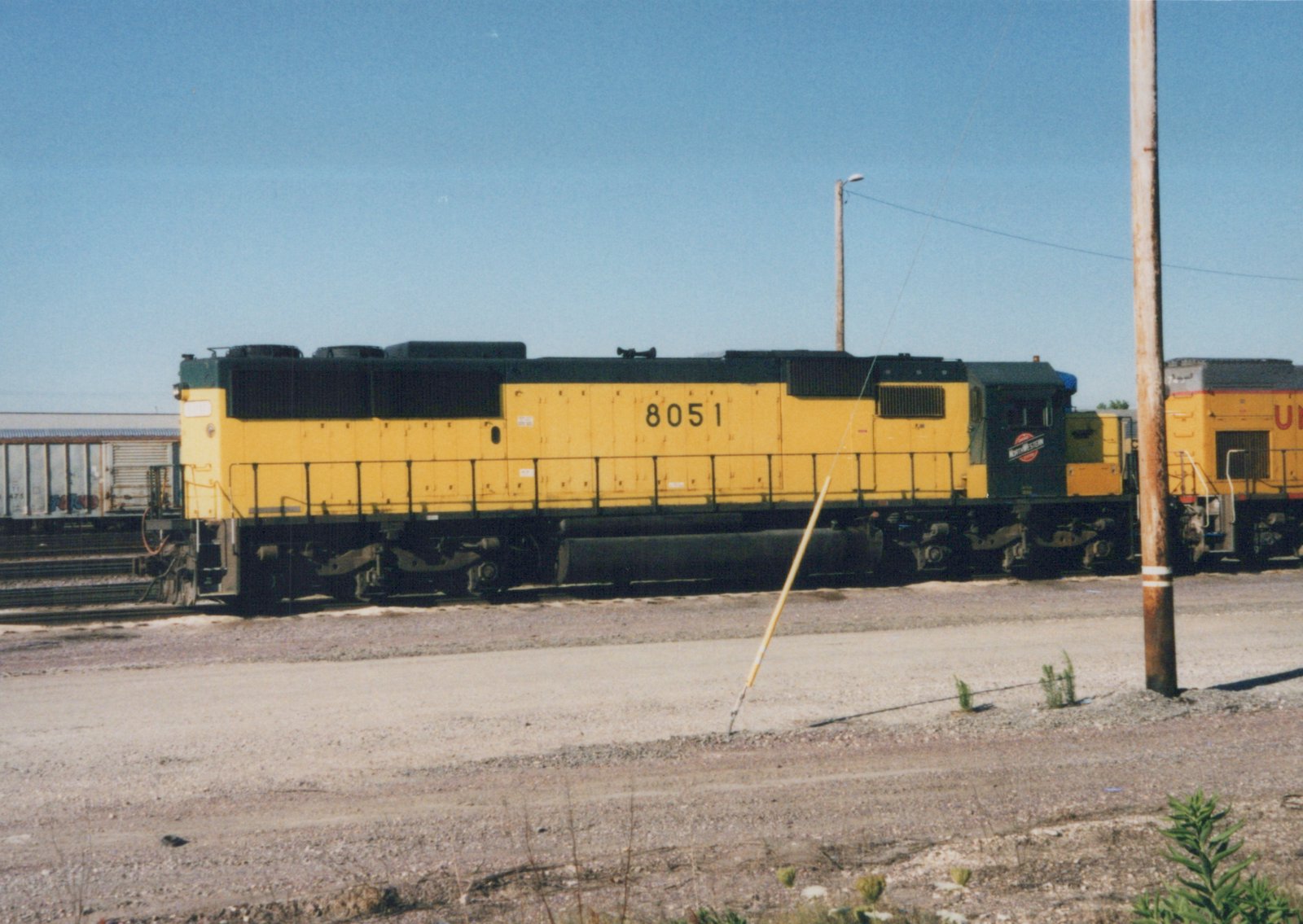 A side view of a yellow and black Chicago and North Western diesel locomotive numbered 8051, parked on tracks in a gravel yard under a clear blue sky.