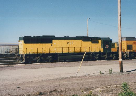A side view of a yellow and black Chicago and North Western diesel locomotive numbered 8051, parked on tracks in a gravel yard under a clear blue sky.