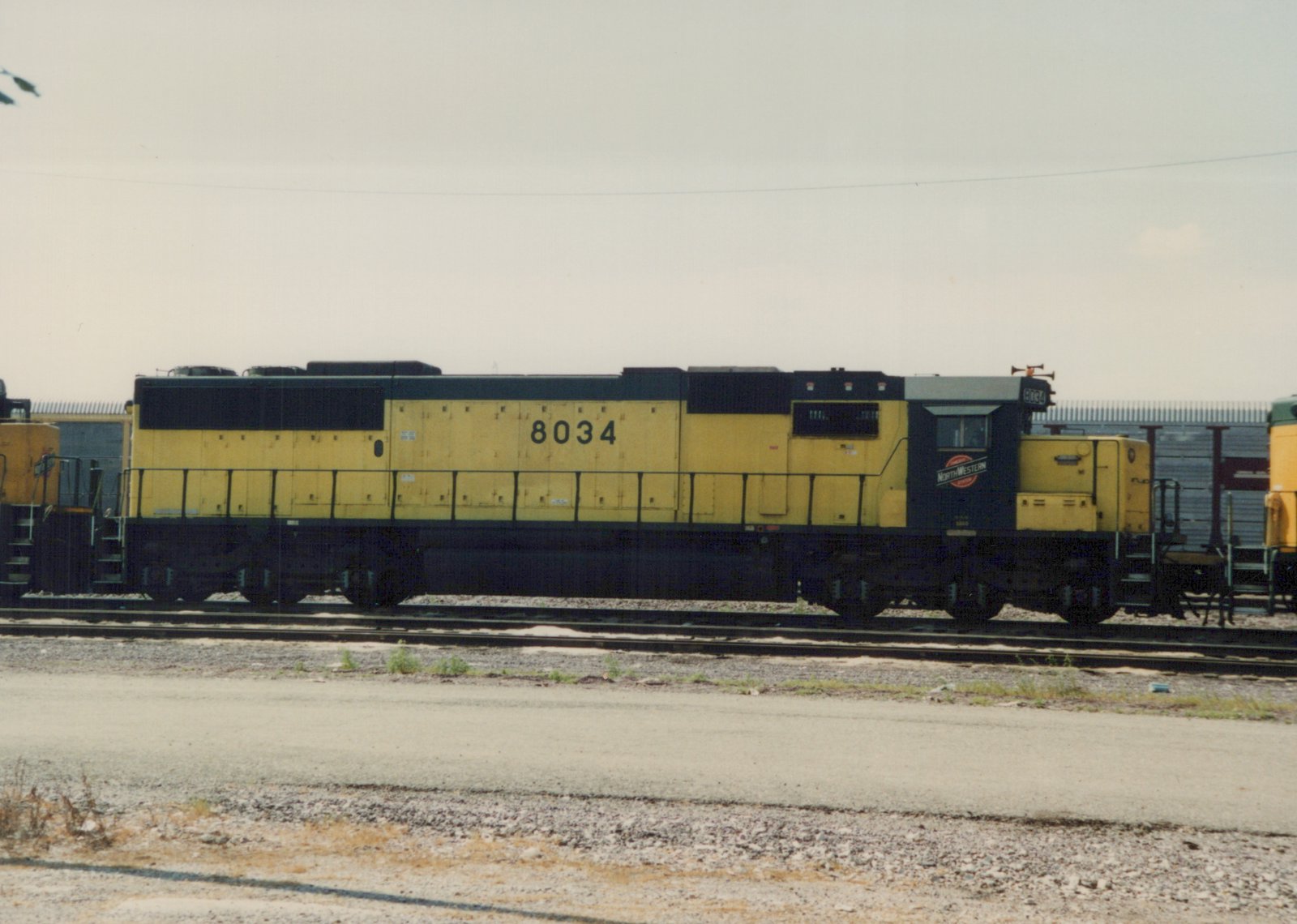 A yellow and black Chicago and North Western diesel locomotive, number 8034, is parked on tracks in a yard under an overcast sky.