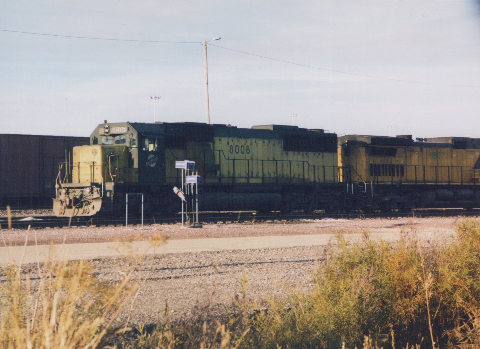 Two yellow and black diesel locomotives, numbered 8008 and 8617 and bearing the Chicago and North Western livery, are parked on tracks in a gravel yard with tall grass in the foreground and a clear sky above.