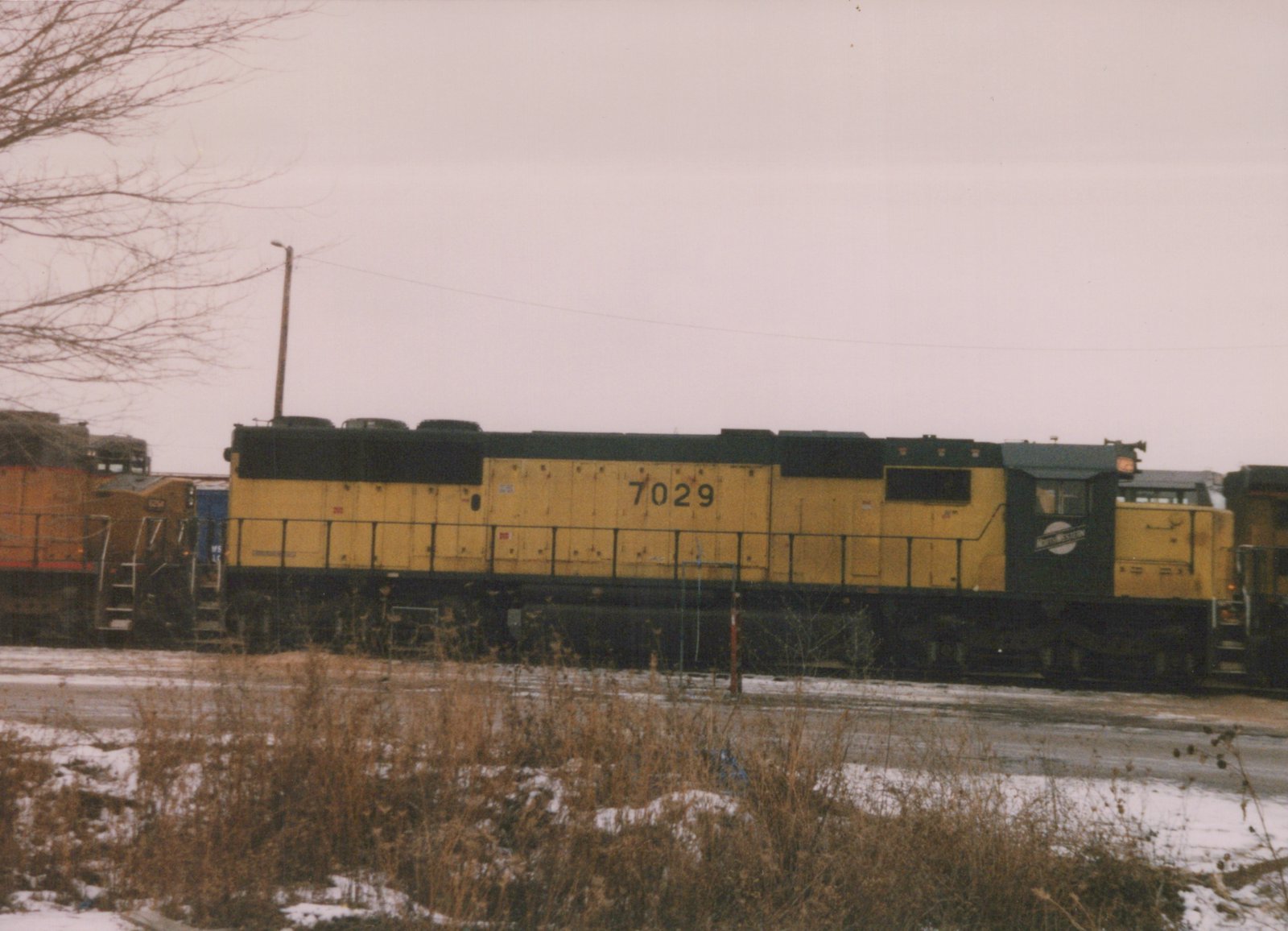 A yellow and black diesel locomotive with the number 7029 and 'Northwestern' lettering on its cab, sitting on tracks in a snow-dusted rail yard under an overcast sky.