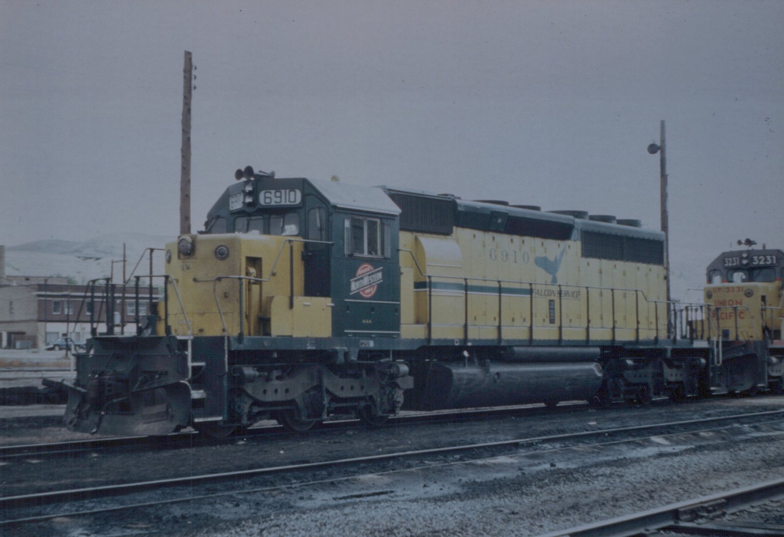 A yellow and green Chicago and North Western locomotive numbered 6910 sits on tracks in a snowy yard, with a Union Pacific locomotive numbered 3231 in the background.