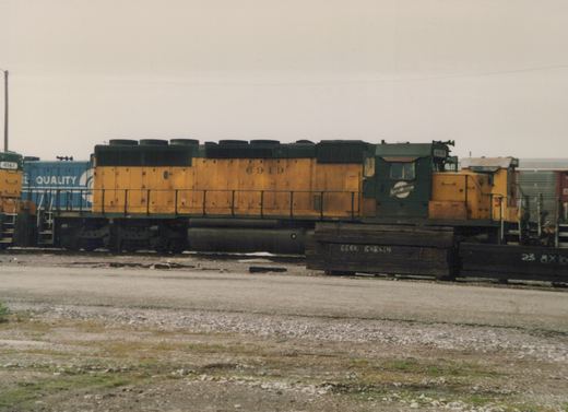 A yellow and dark green Chicago and North Western locomotive, number 6919, stands on a track in a gravel yard with other railcars nearby.