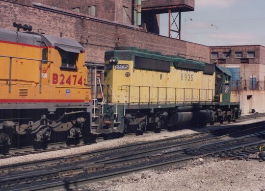 Two diesel locomotives, one green and yellow SD24 numbered 6935 and one yellow and gray GP38-2 numbered B2474, both with Burlington Northern lettering, are coupled together on tracks at a rail yard with brick buildings in the background.