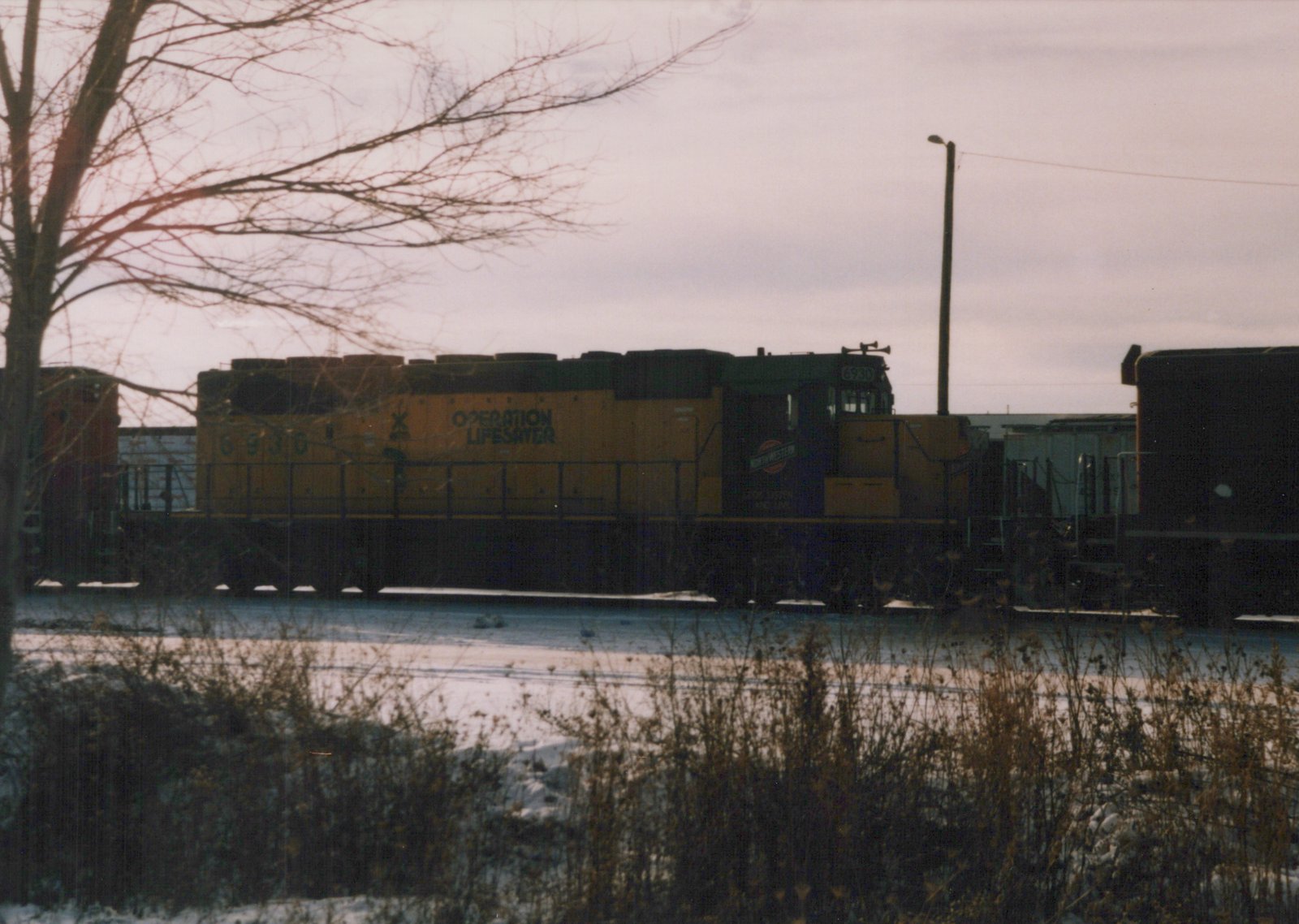 A yellow and green diesel locomotive with the number 6930 and 'Operation Lifesaver' lettering is parked in a snow-covered rail yard with bare trees and other freight cars nearby.