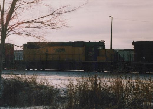 A yellow and green diesel locomotive with the number 6930 and 'Operation Lifesaver' lettering is parked in a snow-covered rail yard with bare trees and other freight cars nearby.