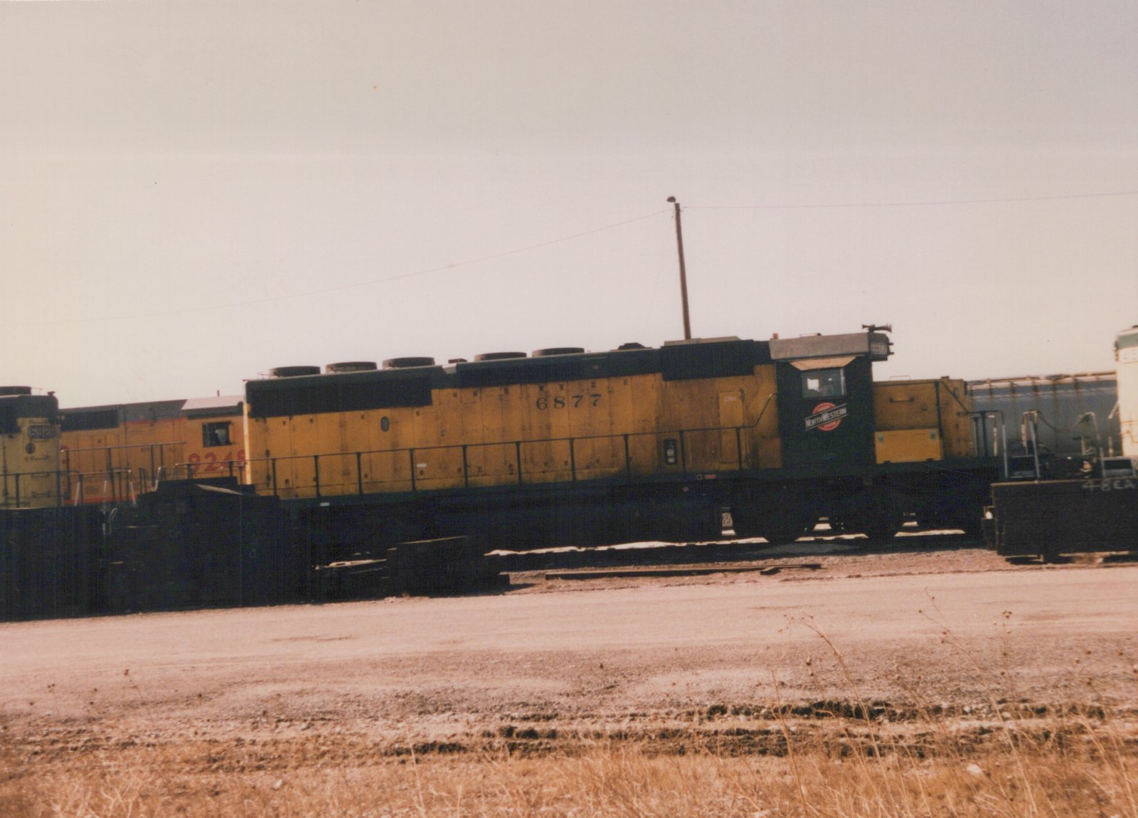 A side view of a yellow and black Chicago and North Western diesel locomotive, number 6877, in a rail yard, with another yellow locomotive behind it and dry ground in the foreground.