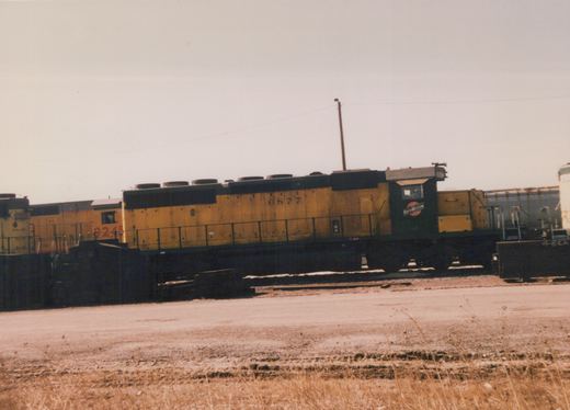 A side view of a yellow and black Chicago and North Western diesel locomotive, number 6877, in a rail yard, with another yellow locomotive behind it and dry ground in the foreground.
