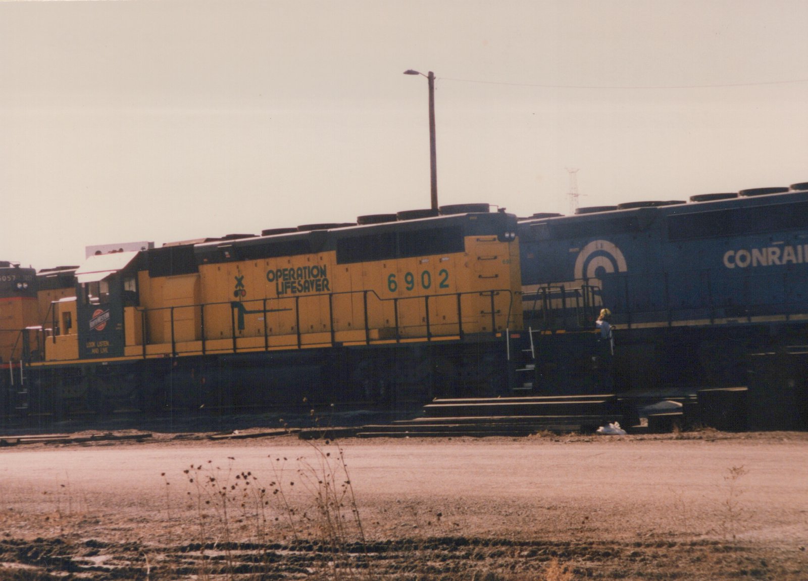 A yellow and black Conrail locomotive numbered 6902 with 'Operation Lifesaver' lettering on its side, standing in a yard with another blue Conrail locomotive behind it.