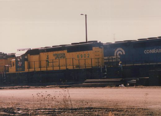 A yellow and black Conrail locomotive numbered 6902 with 'Operation Lifesaver' lettering on its side, standing in a yard with another blue Conrail locomotive behind it.